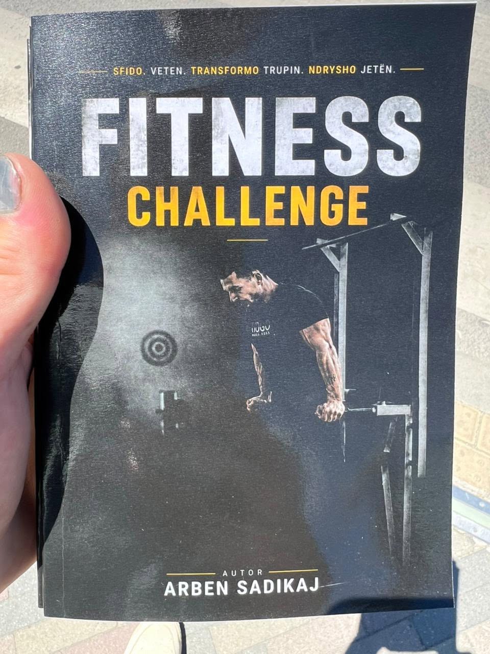 Libri “Fitness Challenge” i autorit Arben Sadikaj, i mbajtur në dorë.
